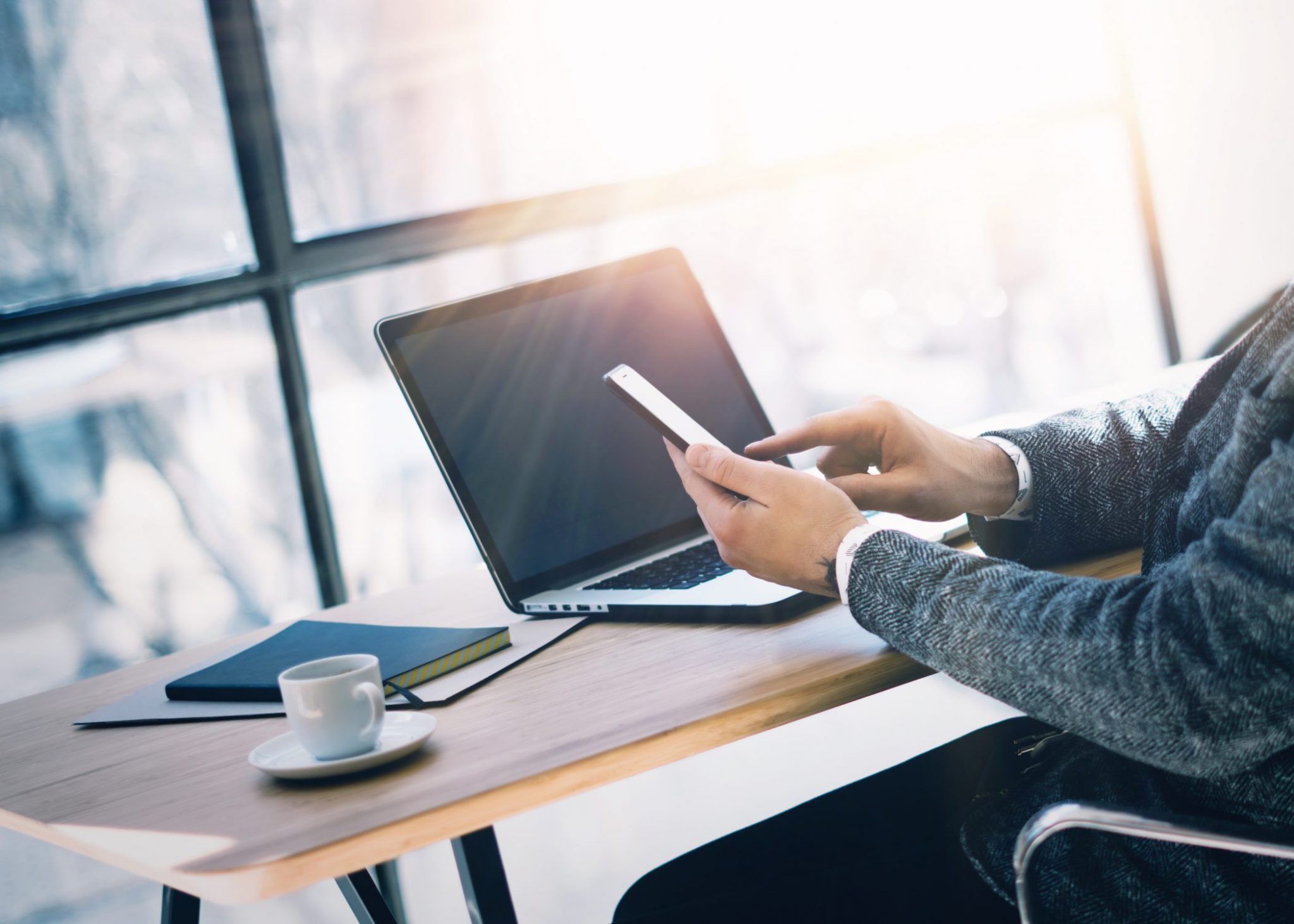 businessman working at the wood table with laptop at sunny office man holding smartphone at hand and texting message horizontal visual effects blurred background e1524505616422